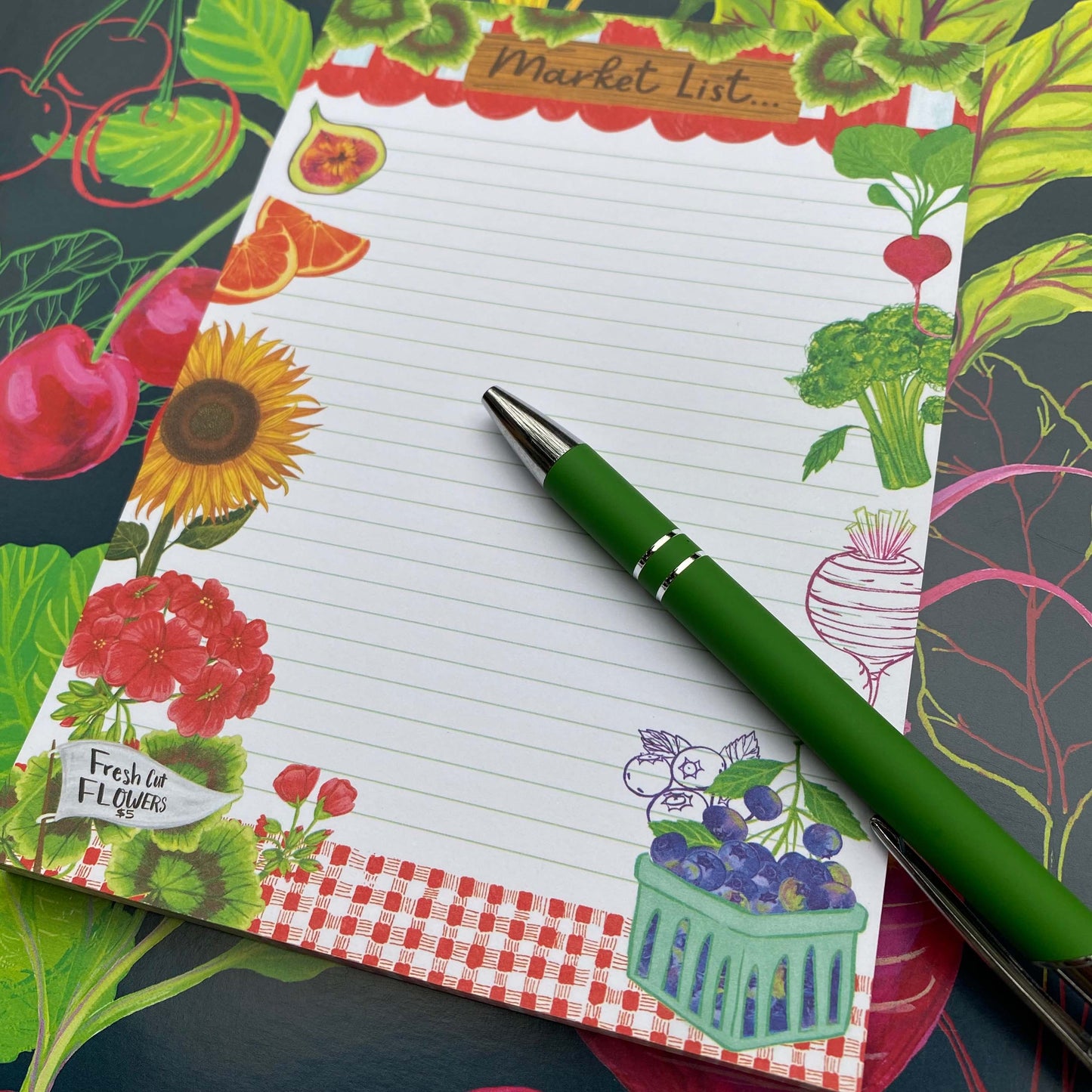 black background with colorful fruits and vegetables. A Market List Notepad sits on top of this illustration containing illustrations of fruits, vegetables and flowers. A green pen sits on top of the notebad.