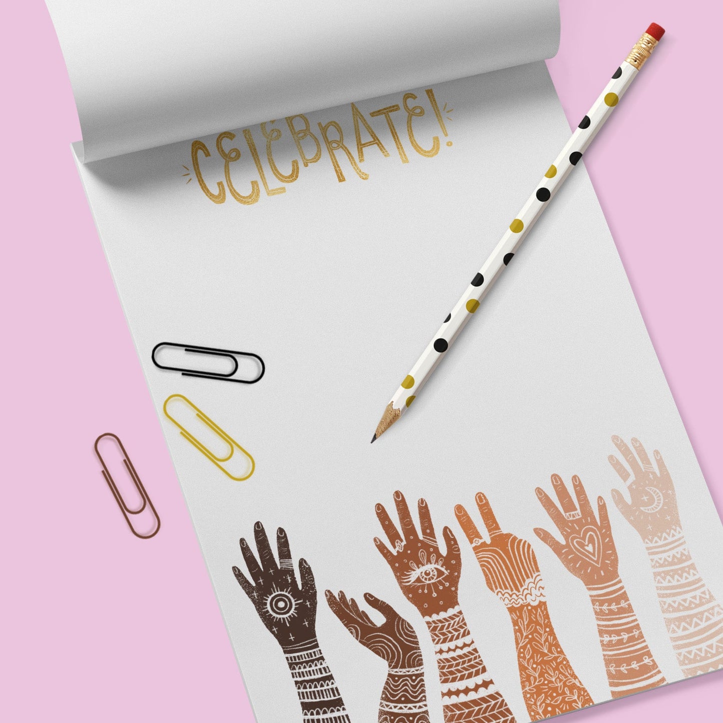 White note pad with diverse multi racially colored hands with arms extended up. the word Celebrate is at the top. There are 3 paperclips ( black, gold and brown) and a white pencil with ochre and black polka dots . These all sit on a lavender colored background.