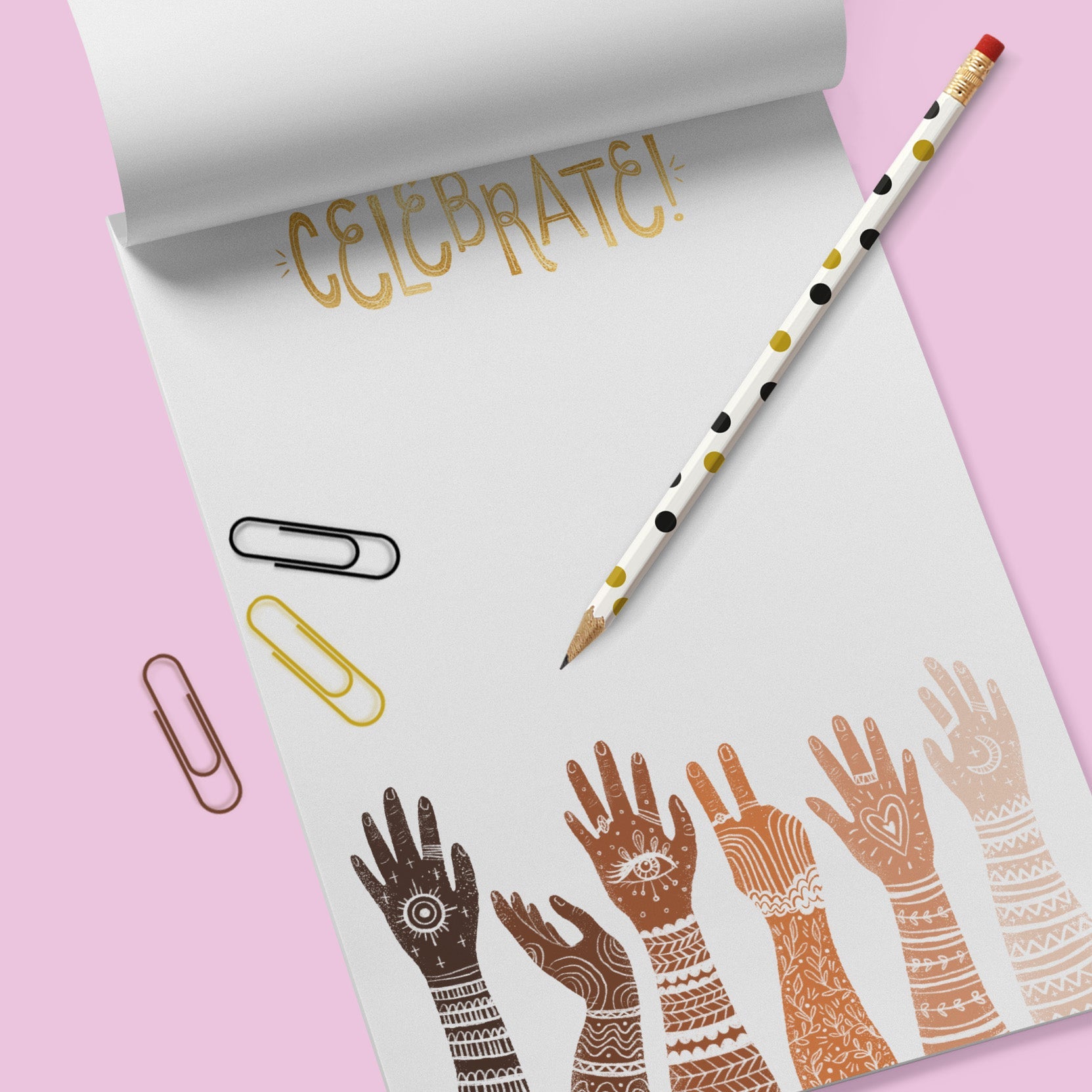 White note pad with diverse multi racially colored hands with arms extended up. the word Celebrate is at the top. There are 3 paperclips ( black, gold and brown) and a white pencil with ochre and black polka dots . These all sit on a lavender colored background. 
