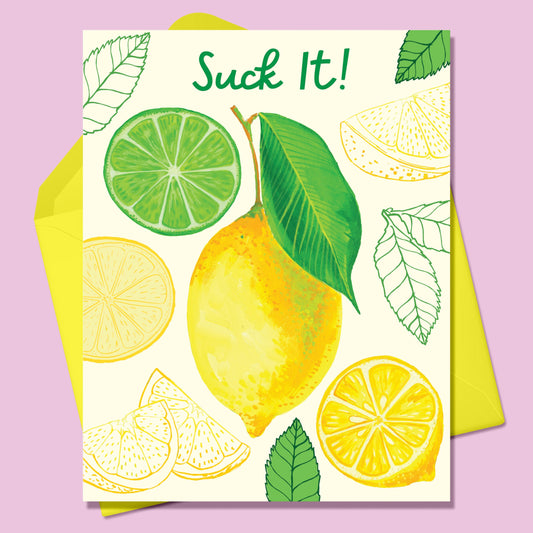 Paper Greeting card with the image of various lemons- a whole lemon in the center surrounded by cut lemons and green leaves and one cut lime  on a cream background. Text says Suck It! With a yellow envelope. On a lavender background.