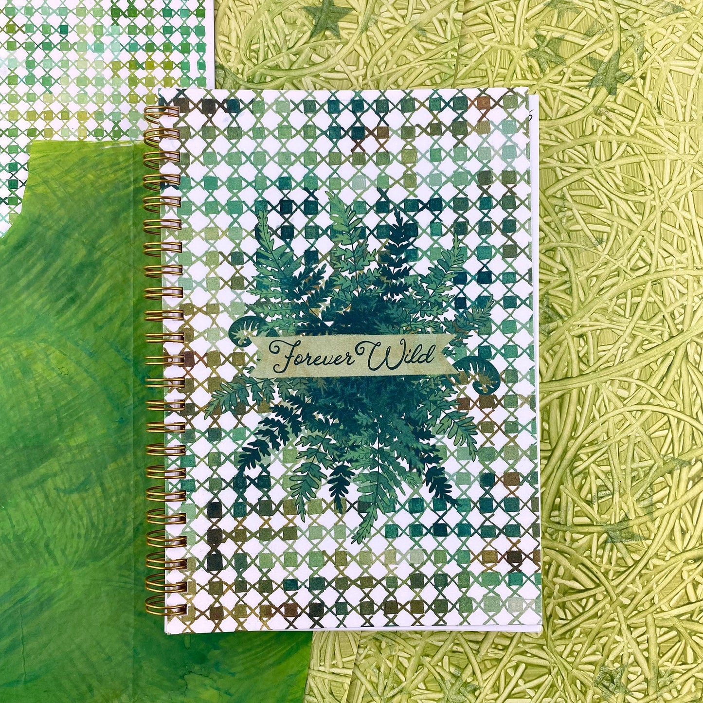 Spiral Bound notebook with a handpainted checkered cover , a grouping of ferns in the center with a banner that says Forever Wild in a hand drawn script font. Very outdoorsy. Using all the green colors. 
