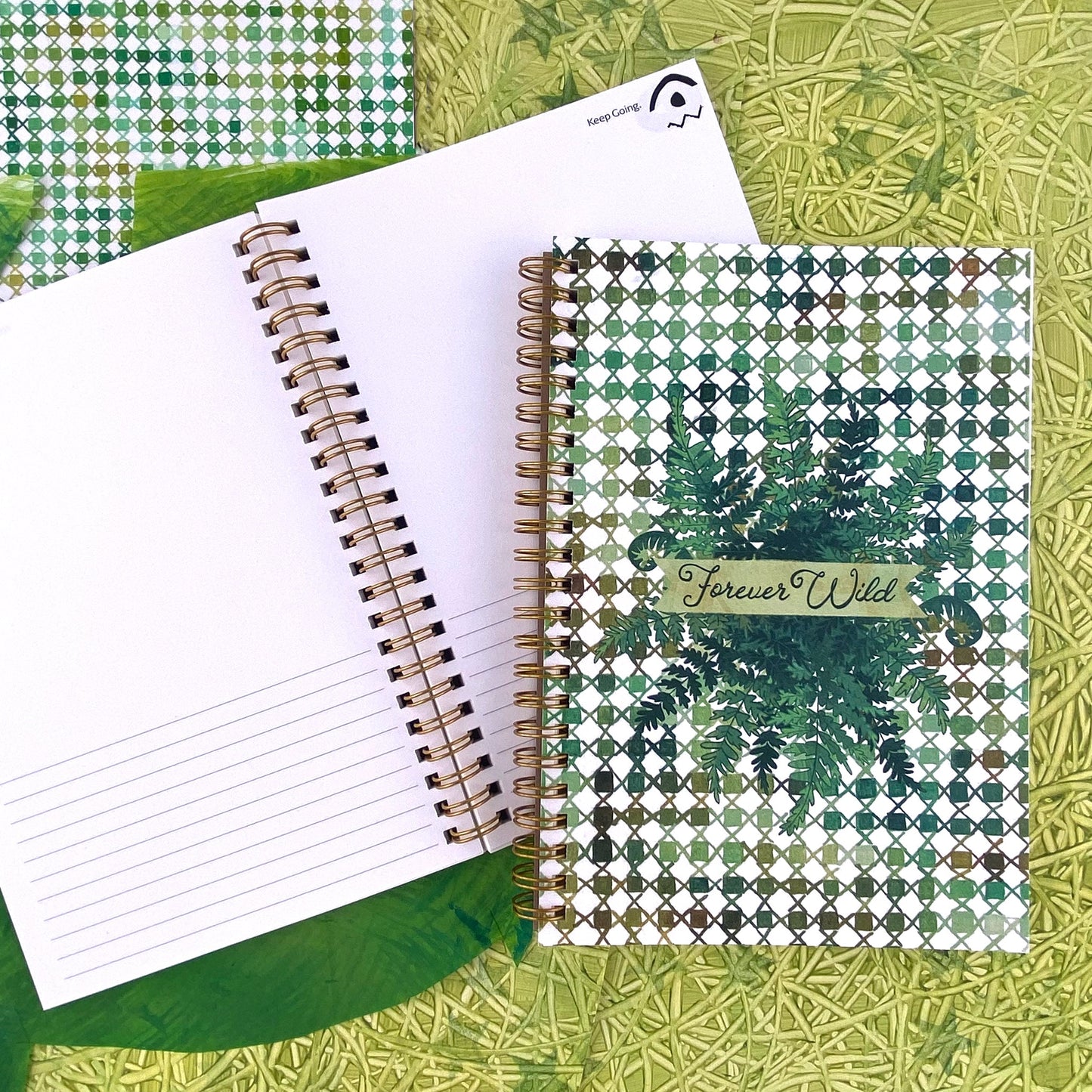 Spiral Bound notebook with a handpainted checkered cover , a grouping of ferns in the center with a banner that says Forever Wild in a hand drawn script font. Very outdoorsy. Using all the green colors. Handpainted green background with swirly textures.
