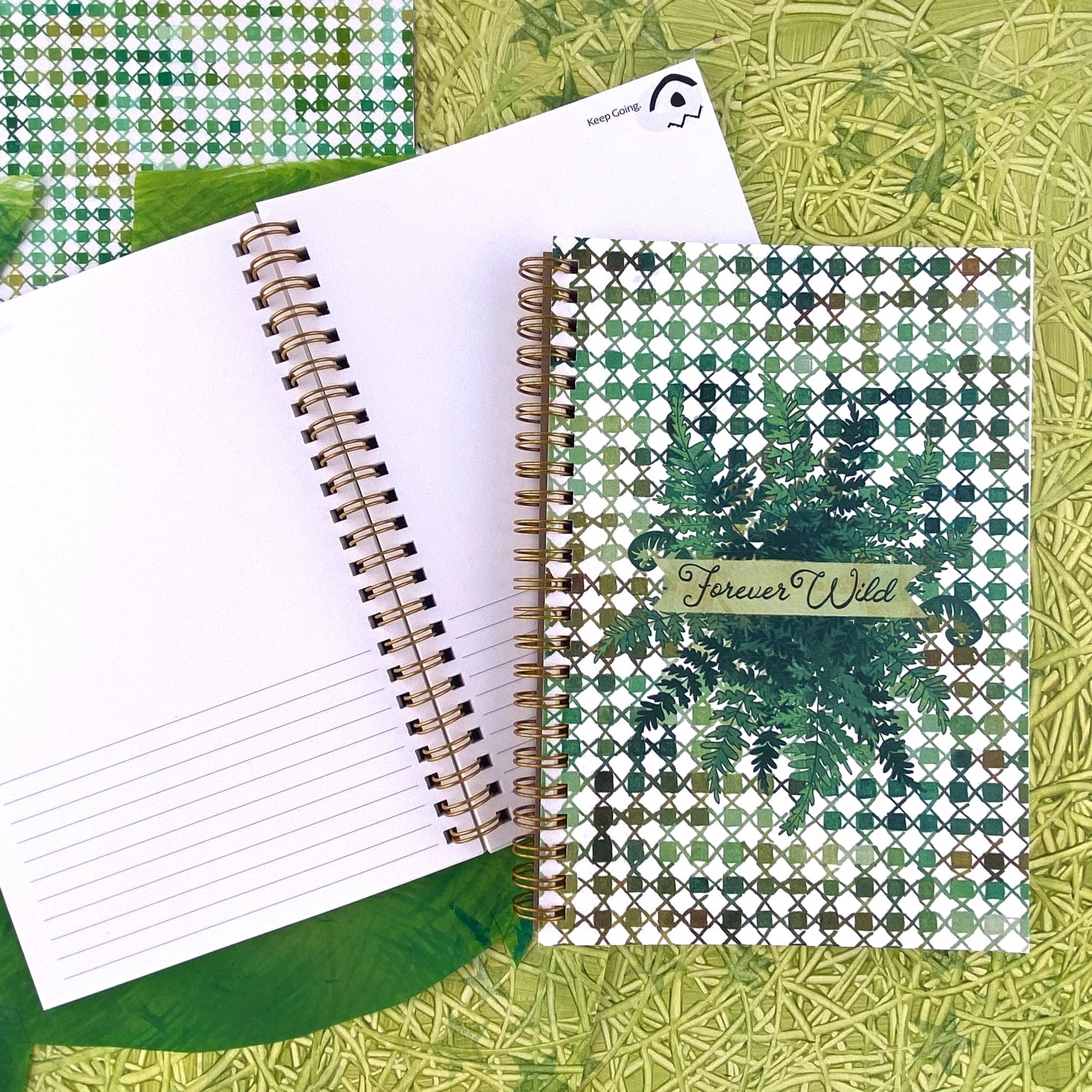 Spiral Bound notebook with a handpainted checkered cover , a grouping of ferns in the center with a banner that says Forever Wild in a hand drawn script font. Very outdoorsy. Using all the green colors. Handpainted green background with swirly textures.