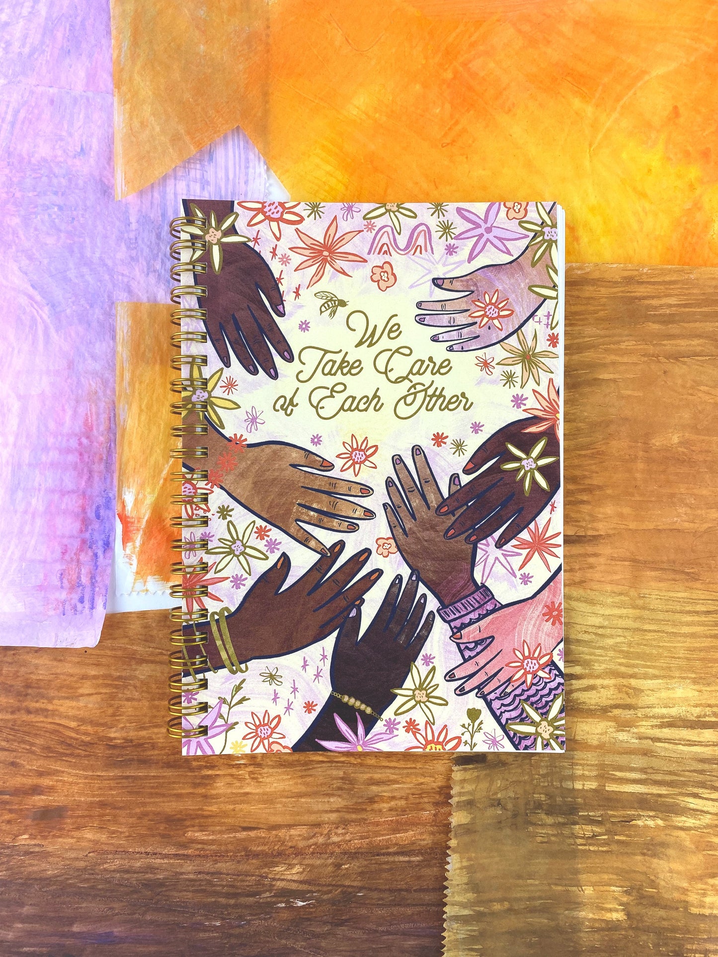 Spiral Bound notebook with hand drawn artwork of multi racial hands and flowers. In the center text says We Take Care of Each Other.